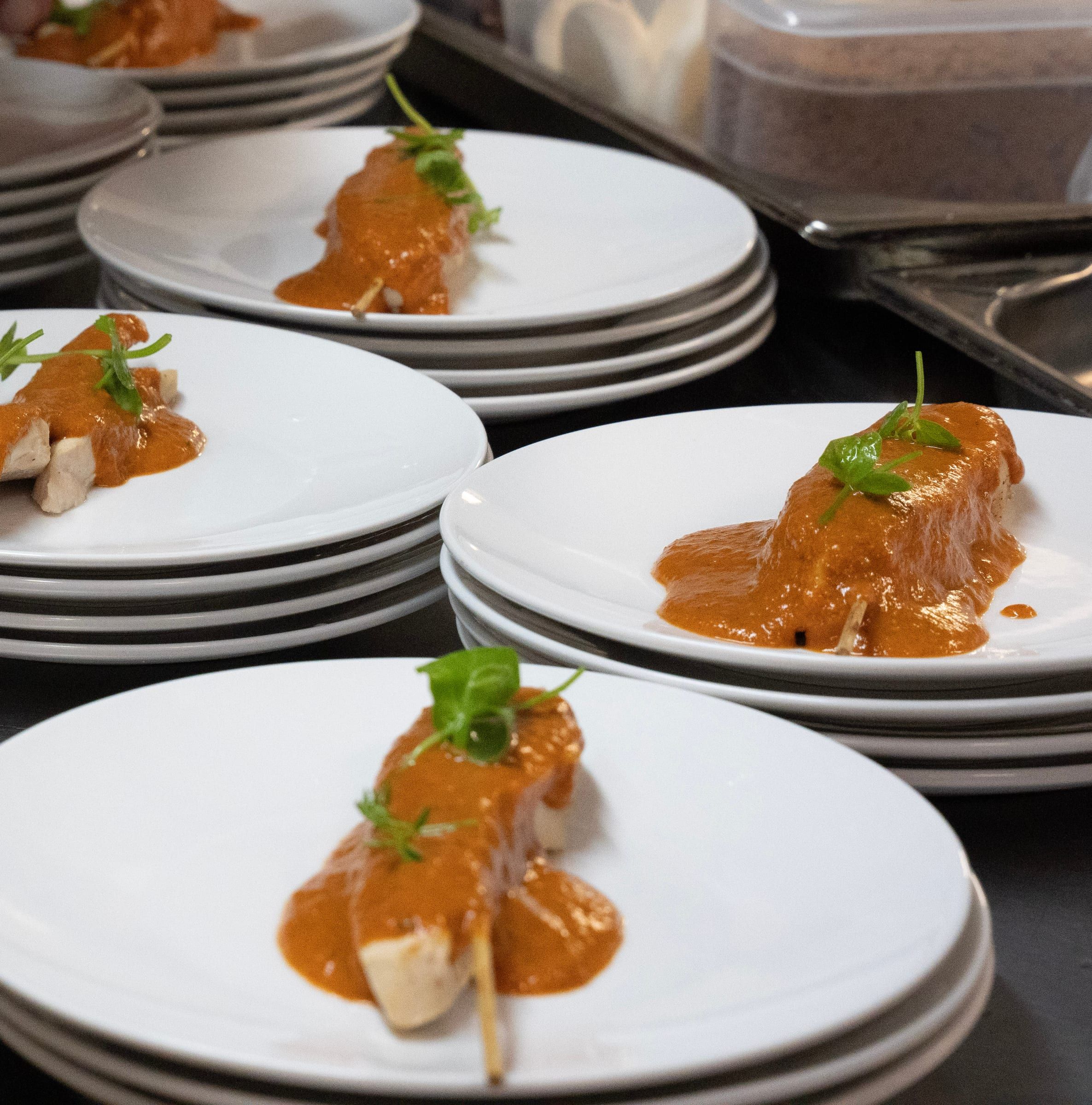 Brochettes de poulet nappées d’une sauce orangée et de jeunes pousses, dressées en série sur des piles d’assiettes blanches.