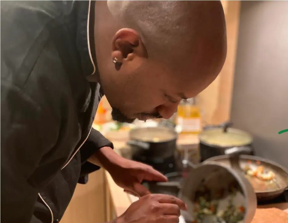 Blaize Iwawe, en veste de chef noire, penché sur ses casseroles en pleine préparation.