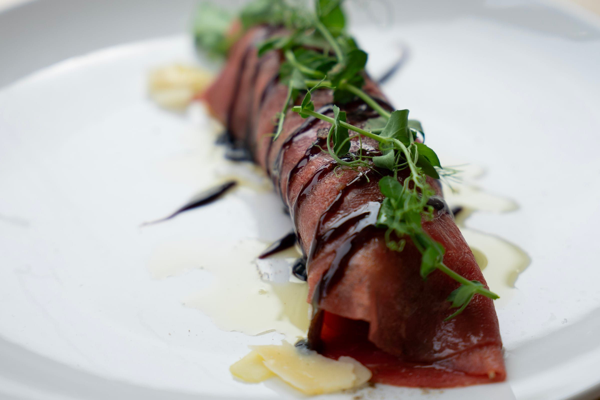 Roulé de carpaccio de bœuf nappé de réduction balsamique, avec des pousses vertes et des copeaux de parmesan.