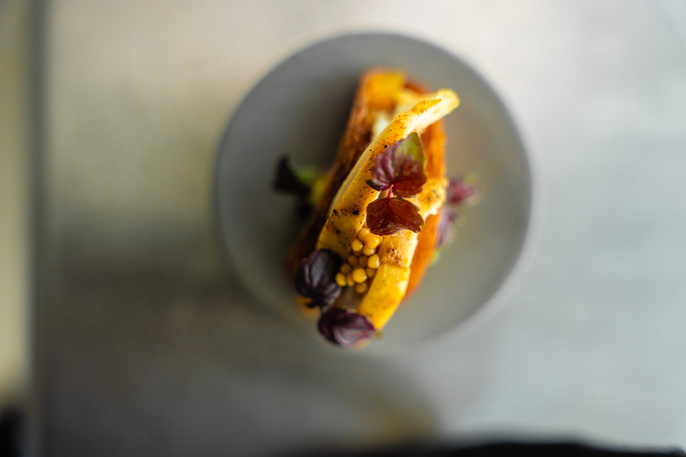 Vue du dessus d’un taco croustillant garni de fines tranches dorées, de petites perles jaunes et de feuilles de shiso pourpre, dans une assiette grise.