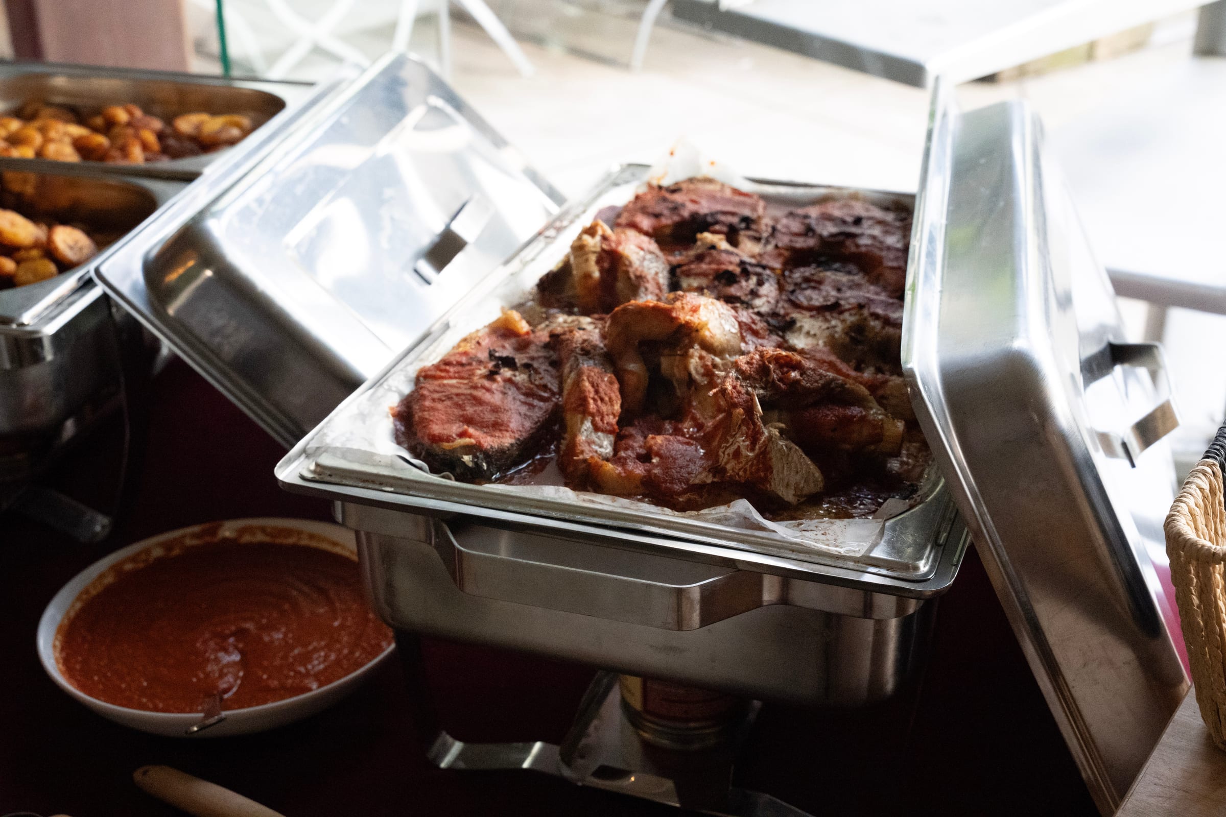 Darnes de poisson grillées dans un chauffe-plat de buffet, avec un bol de sauce rouge et des bananes plantain frites à l’arrière-plan.